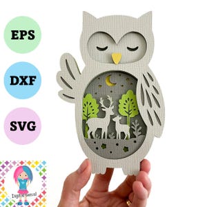 May include: A layered paper owl design with a forest scene inside. The owl is light gray with a yellow beak and closed eyes. The forest scene includes deer, trees, a moon, and stars. The image also includes the text "EPS", "DXF", and "SVG".