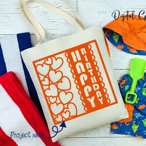 Peut inclure: Un sac fourre-tout en toile blanche avec un motif rouge et blanc qui dit "Joyeux anniversaire" dans un design en forme de c&oelig;ur. Le sac est sur une surface en bois blanc avec une serviette ray&eacute;e bleu et blanc, une serviette ray&eacute;e rouge et blanc, un maillot de bain bleu et orange et un chapeau de soleil orange.