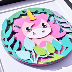 May include: A layered paper art piece featuring a pink unicorn with a yellow horn, surrounded by green and blue foliage. The artwork is framed in a white shadow box, creating a 3D effect. The unicorn has large, expressive eyes.