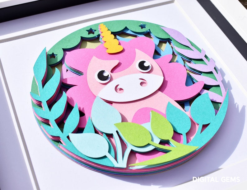 May include: A layered paper art piece featuring a pink unicorn with a yellow horn, surrounded by green and blue foliage. The artwork is framed in a white shadow box, creating a 3D effect. The unicorn has large, expressive eyes.