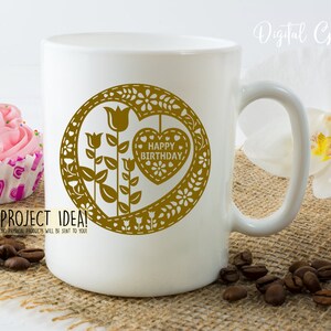 Puede incluir: Taza de cer&aacute;mica blanca con un dise&ntilde;o dorado de un coraz&oacute;n con las palabras "Feliz cumplea&ntilde;os" en su interior. El coraz&oacute;n est&aacute; rodeado de flores y hojas.