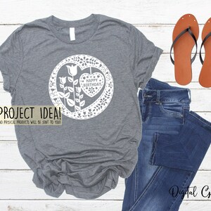 Puede incluir: Una camiseta gris con un dise&ntilde;o blanco de un coraz&oacute;n con las palabras "Feliz cumplea&ntilde;os" dentro de un c&iacute;rculo de flores. El dise&ntilde;o recuerda al corte de papel.