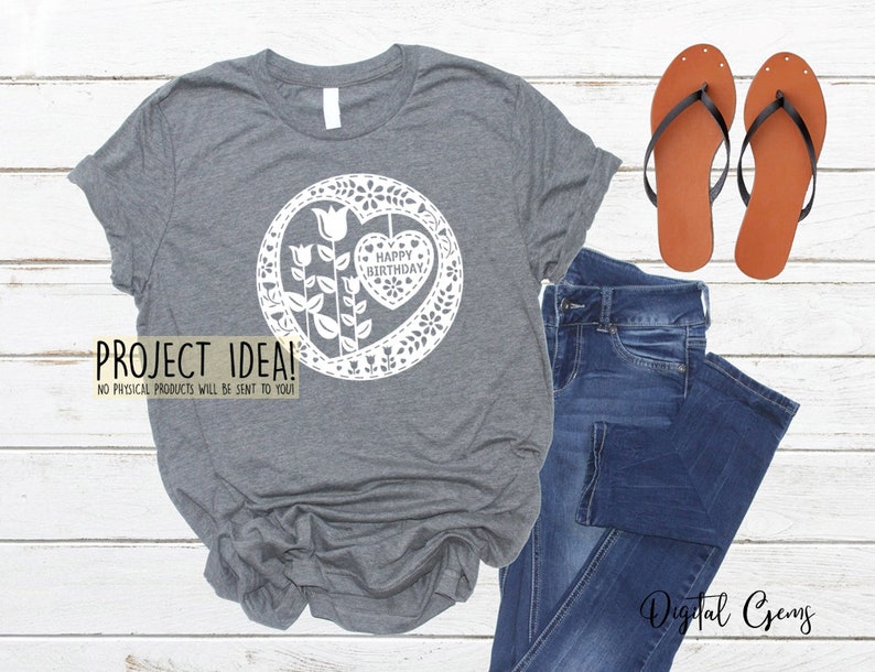 Puede incluir: Una camiseta gris con un dise&ntilde;o blanco de un coraz&oacute;n con las palabras "Feliz cumplea&ntilde;os" dentro de un c&iacute;rculo de flores. El dise&ntilde;o recuerda al corte de papel.