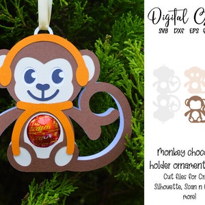 Può includere: Un ornamento di scimmia marrone con orecchie arancioni e una sciarpa, che tiene un dolce di cioccolato. L'ornamento è progettato per essere un porta-dolce di cioccolato. Il testo "Digital Gems SVG DXF EPS FILES" è nell'angolo in alto a destra dell'immagine. Il testo "Monkey chocolate holder ornament design Cut files for Cricut, Silhouette, Scan n Cut and more!" è nell'angolo in basso a destra dell'immagine.