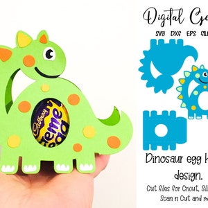 May include: A green dinosaur-shaped egg holder with orange and yellow accents, holding a Cadbury Creme Egg. The design includes the text "Digital Gems" and "Dinosaur egg holder design."