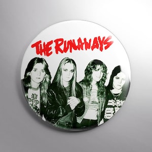 May include: A white button featuring a black and white image of the band The Runaways. The band's name is printed in red, stylized text above the image of the band members.