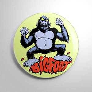 May include: A yellow button with a cartoon illustration of a blue Bigfoot with yellow eyes, standing on its hind legs and raising its fists. The text "BIGFOOT" is written in red letters below the Bigfoot.