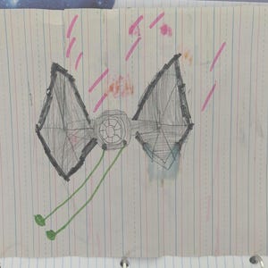 May include: A child's drawing of a TIE fighter from Star Wars, rendered in pencil and crayon on lined paper. The spacecraft is gray with black outlines, green lines, and pink accents. The drawing is centered on the page.