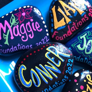 May include: Five painted rocks with names and the words "Foundations 2022" on each. The names are Maggie, Liam, Conner, J, and Kenzie. The rocks are painted in various colors, including black, yellow, blue, green, pink, and red.