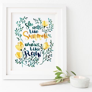 Può includere: Una stampa incorniciata bianca con un'illustrazione ad acquerello di foglie verdi e fiori gialli. Il testo "She acts like Summer + works like Rain" è scritto in un carattere stilizzato.