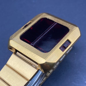 May include: A gold-toned wristwatch with a red digital display and a gold metal band. The watch has a square face and a rectangular display.