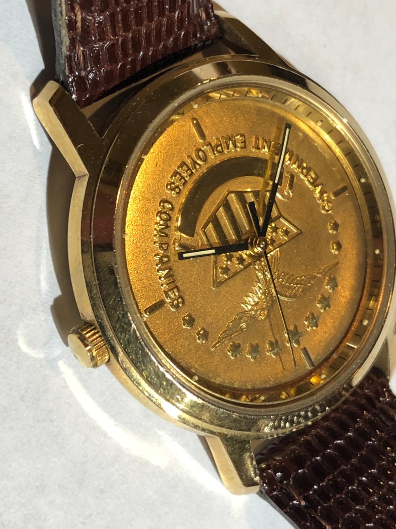 May include: A gold-toned wristwatch with a brown leather strap. The watch face features a gold-coloured design with the text "Government Employees Companies" and a stylised eagle with stars.