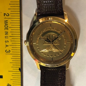 May include: A gold-tone wristwatch with a brown leather strap. The watch face features a gold-coloured emblem with a pyramid, an eagle, and stars. The text around the emblem reads "United States Special Forces Association".