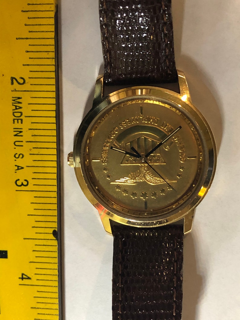 May include: A gold-tone wristwatch with a brown leather strap. The watch face features a gold-coloured emblem with a pyramid, an eagle, and stars. The text around the emblem reads "United States Special Forces Association".