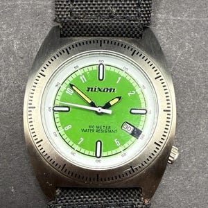 May include: A silver Nixon watch with a green face and black canvas band. The watch face has white markings and the words "100 Meter Water Resistant" and "Nixon".