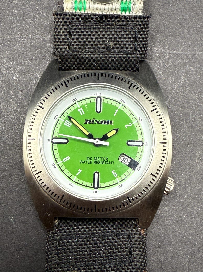 May include: A silver Nixon watch with a green face and black canvas band. The watch face has white markings and the words "100 Meter Water Resistant" and "Nixon".