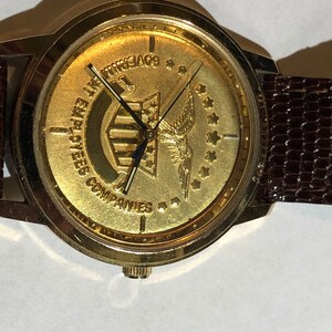 May include: A gold-toned wristwatch with a brown leather strap. The watch face features a gold eagle with a banner that reads "Employees Companies" and "Government 1776".