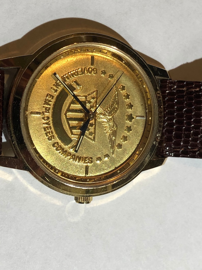 May include: A gold-toned wristwatch with a brown leather strap. The watch face features a gold eagle with a banner that reads "Employees Companies" and "Government 1776".