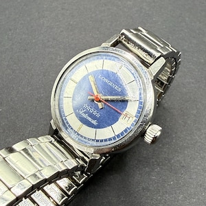 May include: A silver-tone Longines automatic wristwatch with a round blue and silver dial, featuring five stars, a date window at the three o'clock position, a red second hand, and a flexible stainless steel expansion link bracelet.