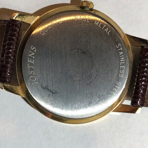 May include: Gold-tone watch with a brown leather strap. The back of the watch has the text "QUARTZ BASE METAL STAINLESS STEEL OSTENS" engraved on it.