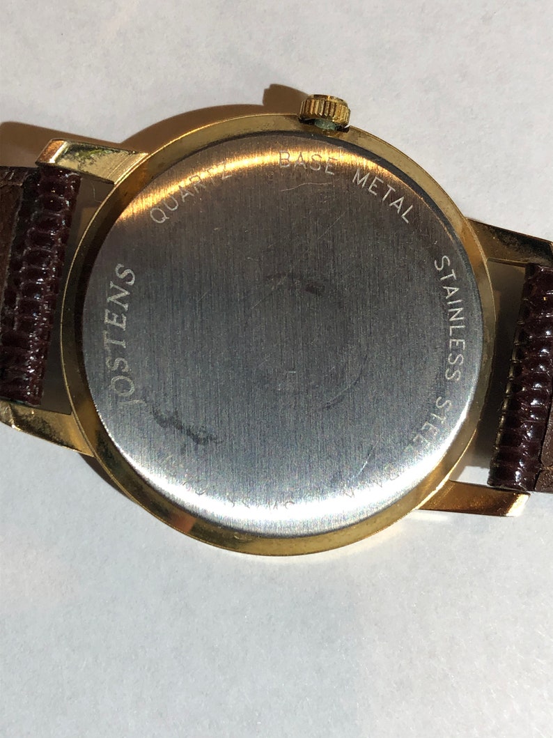 May include: Gold-tone watch with a brown leather strap. The back of the watch has the text "QUARTZ BASE METAL STAINLESS STEEL OSTENS" engraved on it.