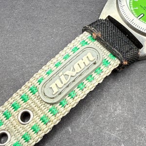 May include: A green and white striped watch band with the word "Nixon" printed on it. The band is made of nylon and has metal eyelets.