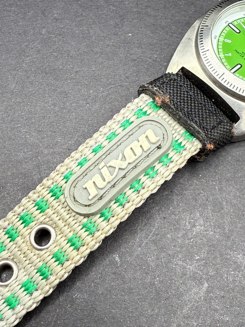 May include: A green and white striped watch band with the word "Nixon" printed on it. The band is made of nylon and has metal eyelets.