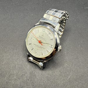 Puede incluir: Un reloj de pulsera Waldman vintage con esfera plateada y correa de acero inoxidable. La esfera del reloj presenta marcadores de hora plateados, un segundero rojo y el texto "Waldman Shockprotected". El reloj está sobre un fondo oscuro.