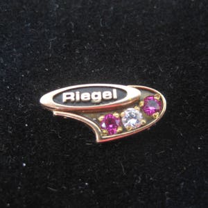 May include: Gold-toned pin with the word "Riegel" in black lettering. The pin has a curved shape and is decorated with pink and white gemstones.