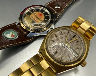 Puede incluir: Dos relojes de pulsera vintage. Uno tiene una esfera dorada y negra con una correa de cuero marrón. El otro reloj tiene una esfera dorada y una pulsera de eslabones dorados. Ambos relojes se muestran sobre una superficie metálica.