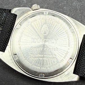 May include: Silver watch with a black nylon strap. The back of the watch has the words "EXACTLY Small Clutch" and "No other watch is closer to the truth." It also says "100 meter Water Resistant Designed in California", "Japan Movement", and "Construction".