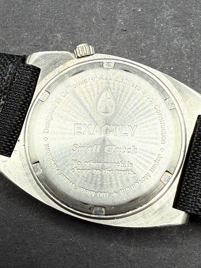 May include: Silver watch with a black nylon strap. The back of the watch has the words "EXACTLY Small Clutch" and "No other watch is closer to the truth." It also says "100 meter Water Resistant Designed in California", "Japan Movement", and "Construction".