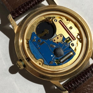 May include: The back of a gold-tone wristwatch with a brown leather strap. The watch face is open, revealing the inner workings of the watch.