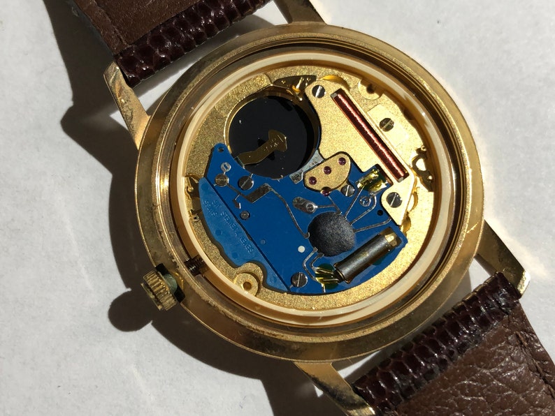 May include: The back of a gold-tone wristwatch with a brown leather strap. The watch face is open, revealing the inner workings of the watch.