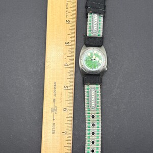 May include: A green and silver wristwatch with a black nylon strap. The strap has the word "JUXON" printed on it in white letters. The watch face has a green background with white markings and a silver bezel.