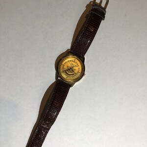 May include: A gold-toned wristwatch with a brown leather strap. The watch face features a gold eagle with a shield and the text "United States of America".