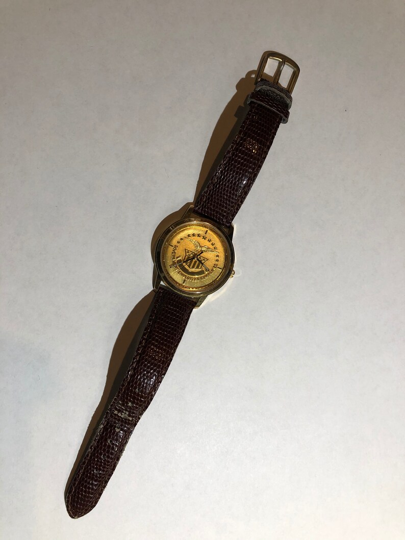 May include: A gold-toned wristwatch with a brown leather strap. The watch face features a gold eagle with a shield and the text "United States of America".