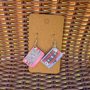 Op de afbeelding: Een paar hangende oorbellen met rechthoekige bedels in de vorm van cassettebandjes. De ene bedel is roze met de tekst RECORD THE BEAUTY, de andere is lichtblauw met de tekst LET'S GET SPOOKY en twee kleine witte spookillustraties.