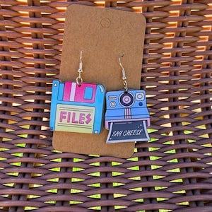 Op de afbeelding: Een paar bungelende oorbellen met een lichtblauwe en gele diskette-bedel met de tekst FILES en een lichtblauwe instant camera-bedel met de tekst SAY CHEESE, beide bevestigd aan zilverkleurige haken op een bruine displaykaart.