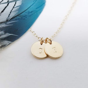 May include: A delicate gold necklace featuring two small, round pendants. Each pendant has a single initial engraved on it. The necklace is set against a white background with a blue and white feather.