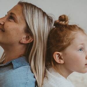 Pode incluir: Um close-up de duas pessoas, uma mulher e uma menina, de costas para a c&acirc;mera. A mulher tem cabelo loiro e est&aacute; vestindo uma camisa listrada azul e branca. A menina tem cabelo ruivo e est&aacute; vestindo uma camisa branca. Ambas est&atilde;o usando pequenos brincos de ouro.