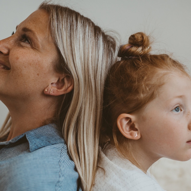 Pode incluir: Um close-up de duas pessoas, uma mulher e uma menina, de costas para a c&acirc;mera. A mulher tem cabelo loiro e est&aacute; vestindo uma camisa listrada azul e branca. A menina tem cabelo ruivo e est&aacute; vestindo uma camisa branca. Ambas est&atilde;o usando pequenos brincos de ouro.