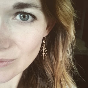 Puede incluir: Primer plano de la cara de una mujer con un pendiente de rama dorada. El pendiente es delicado y presenta un diseño sencillo.