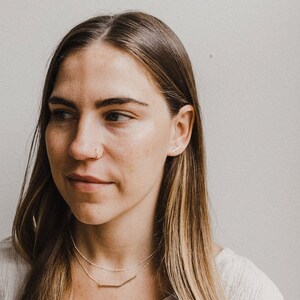 以下が含まれることがあります： 長い茶色の髪をした女性が、銀のネックレスと銀の棒状のイヤリングをしています。白いシャツを着ています。