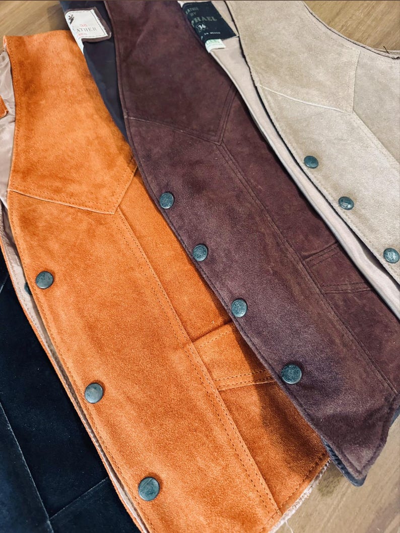 Puede incluir: Primer plano de varios chalecos de ante en tonos naranja, marr&oacute;n y beige. Cada chaleco presenta una parte delantera abotonada y un dise&ntilde;o a medida. Los chalecos se muestran con sus forros interiores visibles.