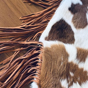 May include: Close-up of a cowhide bag with brown and white patches. The bag features long, tan leather fringe along the edge. The texture of the cowhide is visible, and the fringe adds a rustic, Western-inspired touch.