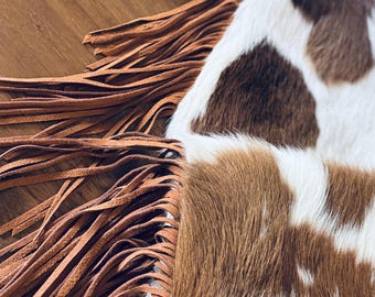 Puede incluir: Primer plano de un bolso de piel de vaca con parches marrones y blancos. El bolso presenta largas y finas tiras de cuero marrón claro en el borde. La textura de la piel de vaca es visible, y las tiras añaden un toque rústico de inspiración occidental.