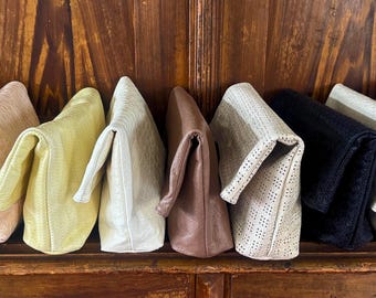 Puede incluir: Una colección de diez bolsos de mano rectangulares con solapa dispuestos en fila. Los bolsos presentan diversas texturas y colores, incluyendo rojo, rosa claro, amarillo, blanco, marrón, blanco perforado, negro, crema y bronceado, con un diseño simple.