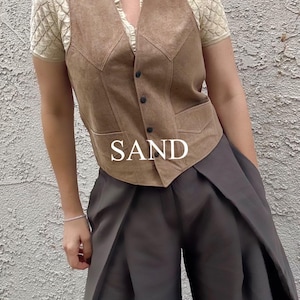 Puede incluir: Un chaleco de ante color arena con cierre de botones, usado sobre una blusa crema con mangas texturizadas. El chaleco se combina con pantalones de pierna ancha de color gris oscuro. La palabra "SAND" se muestra en texto blanco.