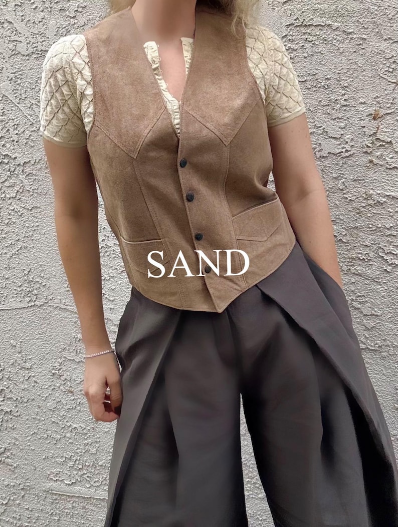 Puede incluir: Un chaleco de ante color arena con cierre de botones, usado sobre una blusa crema con mangas texturizadas. El chaleco se combina con pantalones de pierna ancha de color gris oscuro. La palabra "SAND" se muestra en texto blanco.